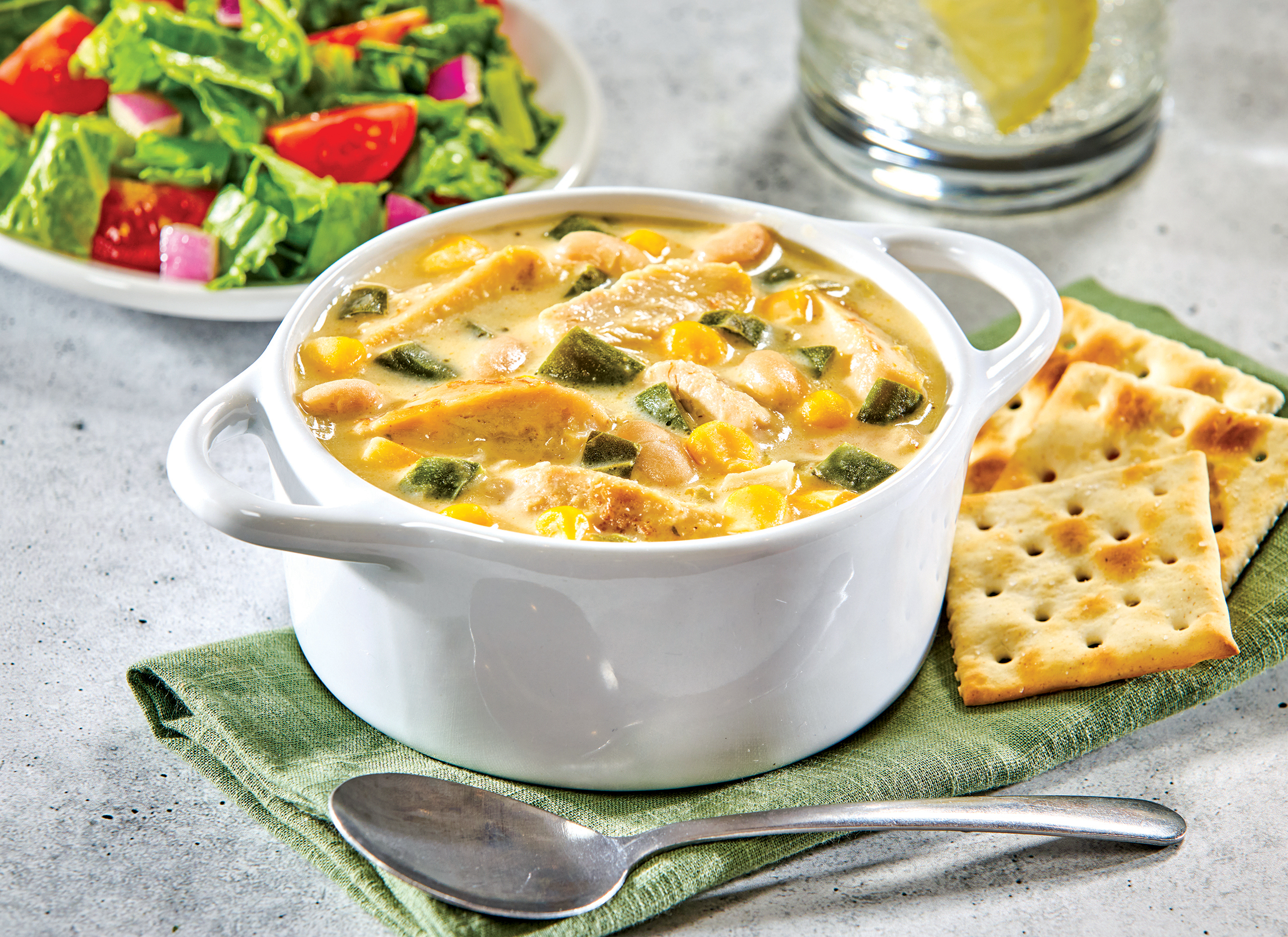 A bowl of creamy soup with chicken, beans, corn, and green peppers sits on a green napkin. Crackers, a spoon, a salad, and a glass of water with lemon are also on the table.