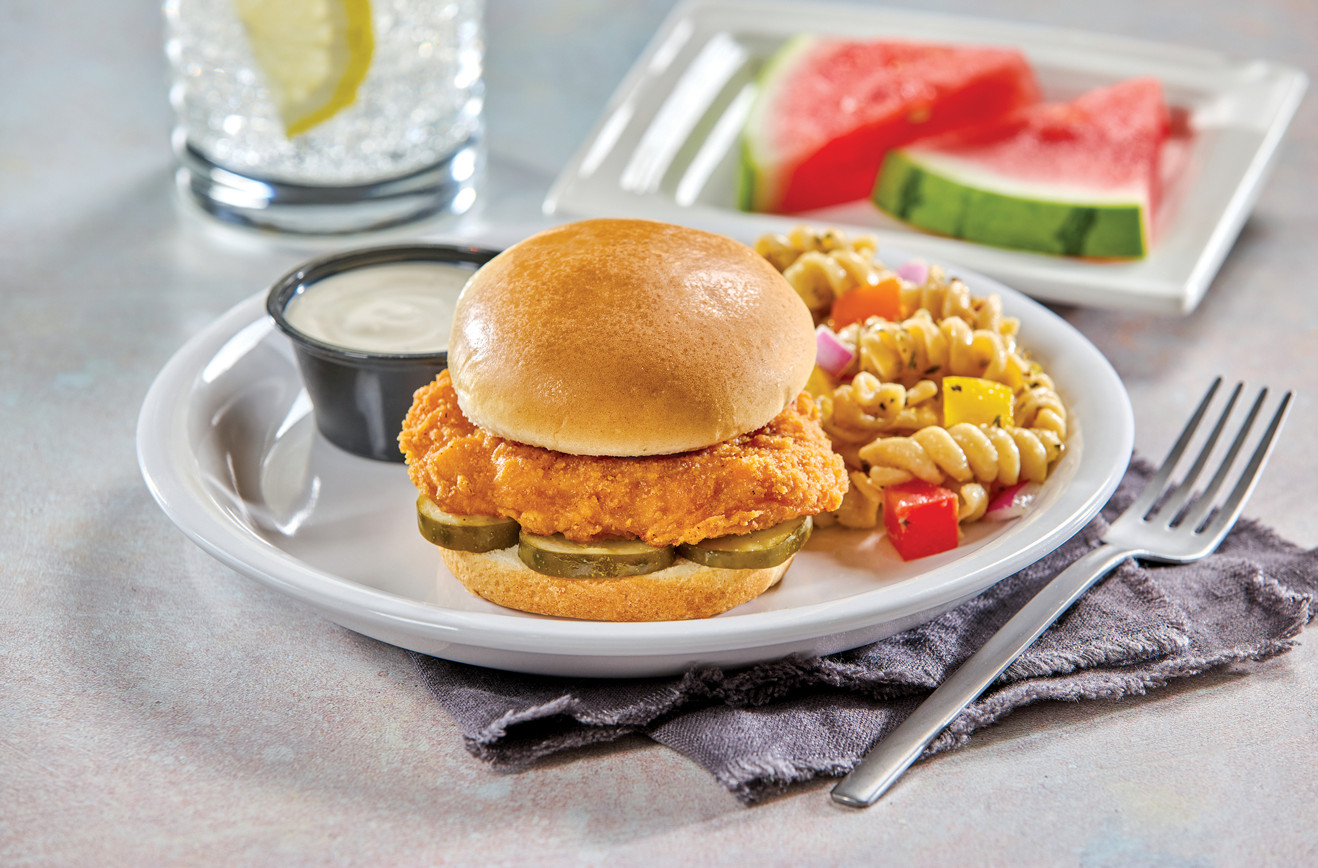 A plate with a fried chicken sandwich on a bun with pickles, served with pasta salad and a cup of ranch dressing. In the background, there are watermelon slices and a glass of water with lemon.