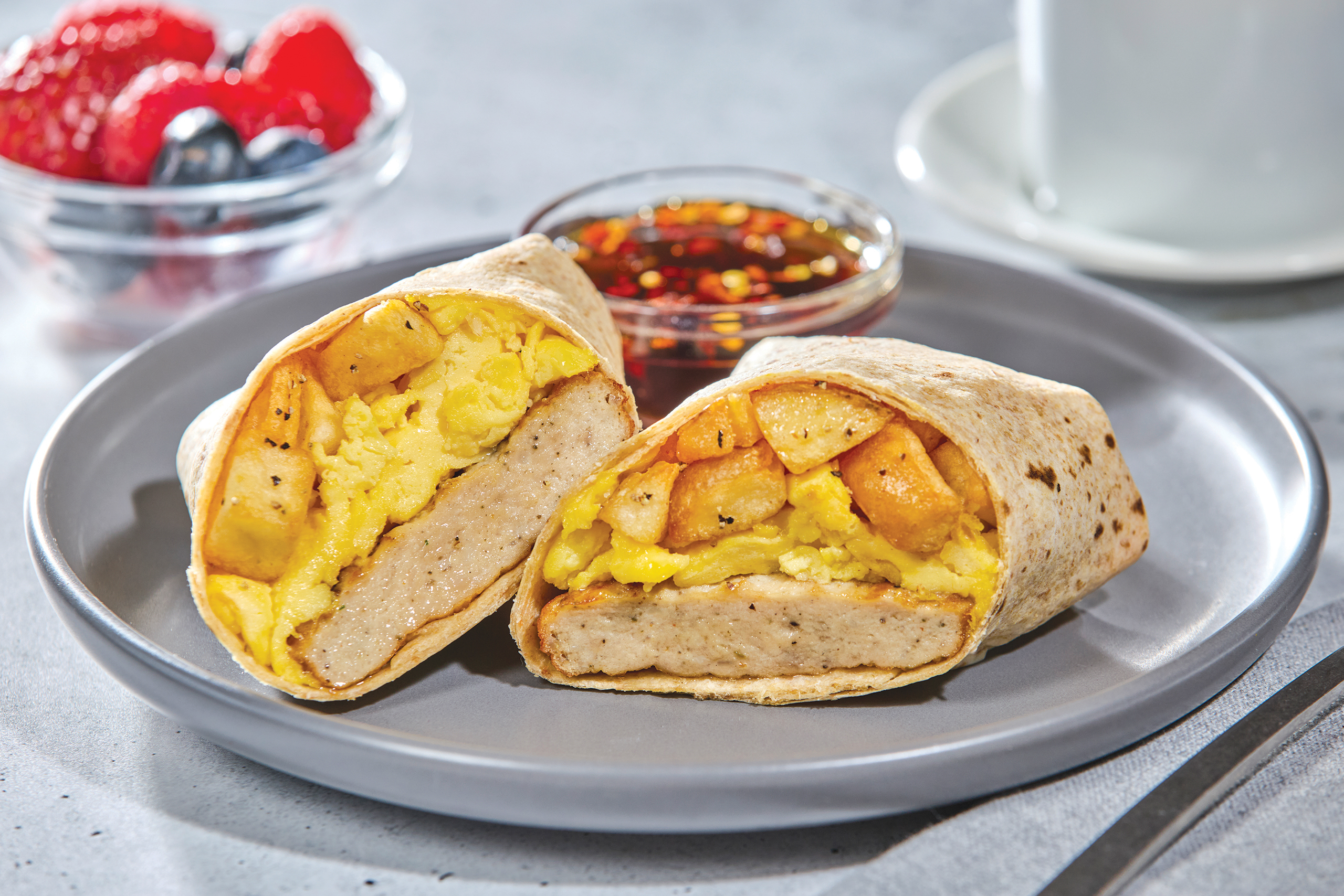 A breakfast burrito cut in half on a gray plate, filled with eggs, potatoes, and a chicken patty. A small bowl of sauce, a cup, and a dish of mixed berries are in the background.