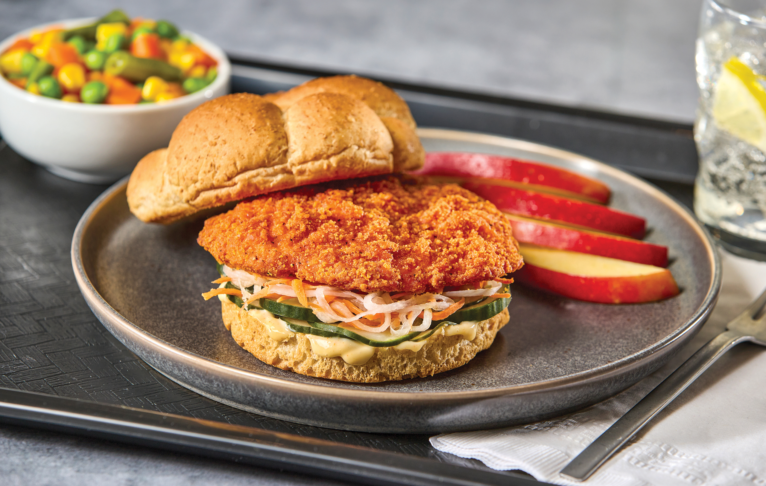A breaded chicken sandwich with vegetables on a bun, served with apple slices and a bowl of mixed vegetables on the side. A glass of water with lemon is in the background.